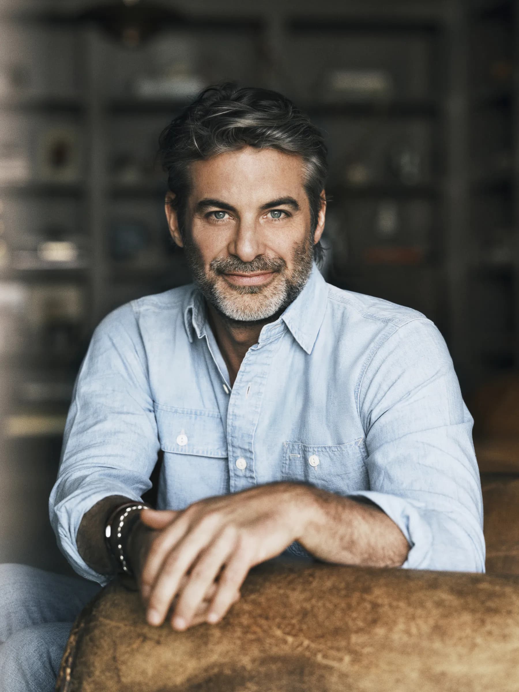 Jim Curtis in a chambray shirt, resting his arm on a leather couch and looking straight at the camera